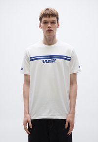 Bela bombažna majica s svetlo modro vodoravno črto in logotipom "CALVIN KLEIN" na prsih. Elastičen ovratnik in kratki rokavi.