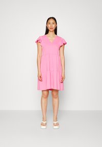 Vestido de algodão rosa com decote em V, mangas curtas em babado, design de saia em camadas e um corte confortável. Usado com sandálias brancas de salto grosso.