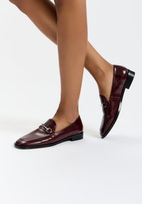 Burgundské kožené mokasíny se čtveratým špičkou, nízkým černým podpatkem a stříbrnou sponou na nártu. Hladká textura a elegantní design.