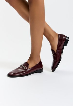 Mocassins en cuir bordeaux avec un bout carré, un petit talon noir et une boucle argentée sur l'empeigne. Texture lisse et design épuré.