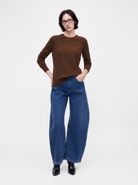 Femme aux cheveux courts et foncés avec des lunettes, portant un haut marron à manches longues, un jean bleu à jambes larges et des bottines noires, debout les mains dans les poches.