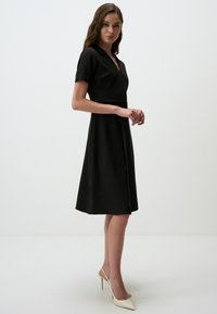 Schwarzes geripptes Kleid mit V-Ausschnitt und kurzen Ärmeln, ausgestelltem Rock und taillierter Taille. Getragen mit cremefarbenen spitzen Absatzschuhen.