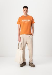 T-shirt laranja de algodão com texto branco, calças pregueadas bege e botas castanhas. A segurar uma bolsa de lona creme. Fundo minimalista.