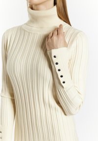 Kermanvärinen ribbipaita, jossa on korkea turtleneck-kaulus ja napitus hihansuissa. Pehmeä tekstuuri ja vartalonmyötäinen leikkaus.