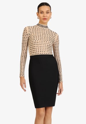 Frau trägt ein beigefarbenes langärmeliges Oberteil mit schwarzen Quadratmustern und einen eng anliegenden schwarzen knielangen Rock, steht vor einem weißen Hintergrund.