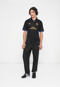 Μαύρη ζακέτα με μπλε λεπτομέρειες και χρυσό κείμενο "TRUE COLOURS." Συνδυασμένη με μαύρο παντελόνι και μαύρα υποδήματα με λευκές λεπτομέρειες.