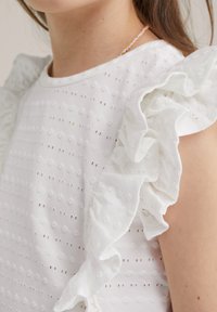 Bílý top s volánovými rukávy, texturovaná látka s tečkovaným vzorem a horizontálními pruhy. Detailní pohled zdůrazňující designové detaily.