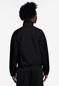 Casaco corta-vento preto com gola alta, cintura elástica e costura contrastante em padrão de onda nas costas.