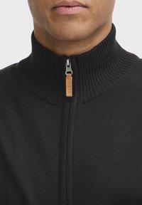 Sort zippered sweater med en høj ribbet krave. Har en sølv lynlås og et læderdetalje med ordet "BLEND" indgraveret. Glat tekstur.