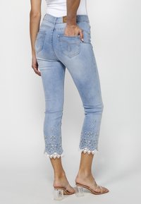 Light blue denim jeans with embroidery along the hem, featuring a scalloped lace detail. Two back pockets with decorative stitching.