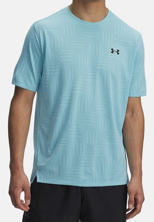 Mężczyzna ubrany w jasnoniebieską sportową koszulkę z krótkim rękawem Under Armour z subtelnym geometrycznym wzorem i czarne szorty na białym tle.