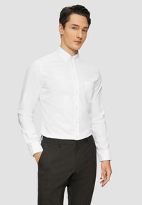 Chemise blanche avec un col boutonné et une poche poitrine, associée à un pantalon sombre. Le tissu semble lisse avec une texture minimale.