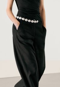 Zwarte wijde broek met hoge taille, gecombineerd met een glanzende zilveren ovale riem. Het bovenstuk is een mouwloze, aansluitende zwarte stof.