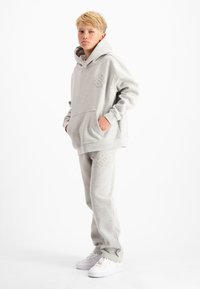 Blonde jongen staat met zijn handen in zijn zakken, gekleed in een grijze hoodie en bijpassende joggingbroek met witte sneakers voor een witte achtergrond.
