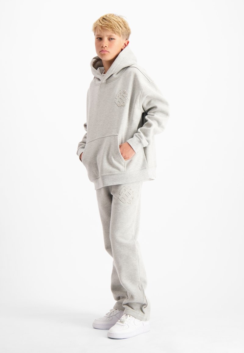 Blonde jongen staat met zijn handen in zijn zakken, gekleed in een grijze hoodie en bijpassende joggingbroek met witte sneakers voor een witte achtergrond.