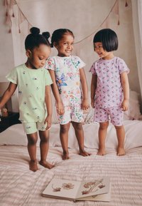Tres niños están de pie sobre una cama, vestidos con pijamas coloridos. El conjunto verde tiene patrones florales, el blanco presenta un diseño de mariposa y el púrpura muestra pequeñas flores.