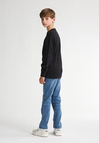 Maglietta nera a maniche lunghe con polsini a coste, abbinata a un paio di jeans in denim e sneakers bianche con logo. Il modello si trova di spalle contro uno sfondo neutro.