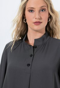 Blonde woman with blue eyes wearing silver hoop earrings and a dark gray buttoned shirt with a mandarin collar against a white background.