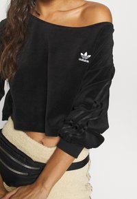 Svart kort sweatshirt i velourtyg, med off-shoulder-design och vitt Adidas-logotyp på bröstet, samt ribbade ärmslut.