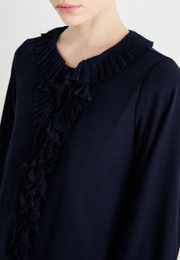 Blusa azul-marinho com um acabamento de ruffles pregueados ao longo do decote, com uma textura suave e um corte relaxado. Os ruffles estendem-se verticalmente pela frente.