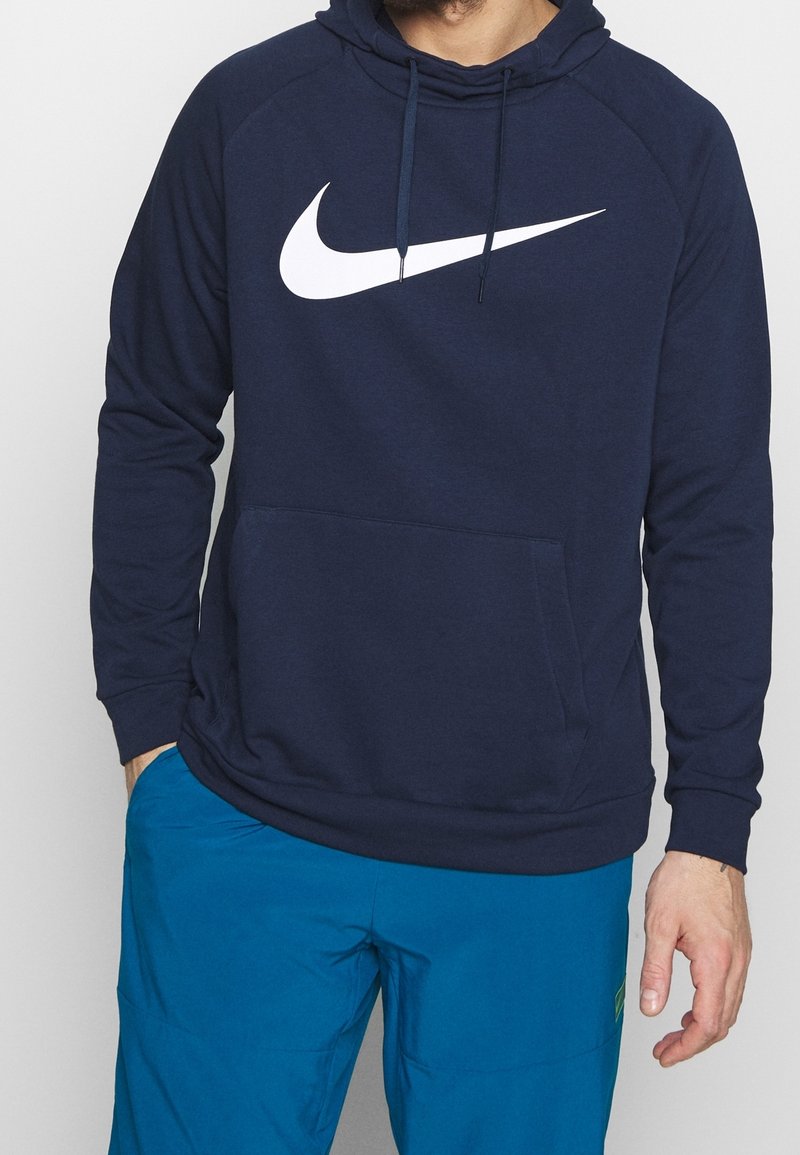 Homme portant un sweat à capuche Nike bleu marine avec logo swoosh blanc et pantalon bleu vif, debout avec une main dans la poche.