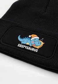 Black knitted beanie with a rectangular patch featuring a blue cartoon dinosaur in a Santa hat, text reading "SLEEPYSAURUS" below.