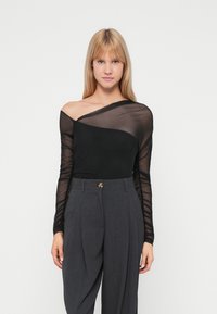 Top lungo a maniche nere in rete con scollatura asimmetrica abbinato a pantaloni grigi a vita alta con pieghe e un bottone decorativo.