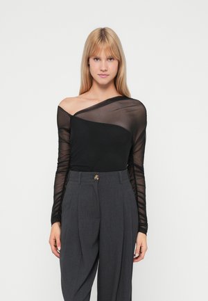 Top en maille noir à manches longues avec un encolure asymétrique, associé à un pantalon gris taille haute avec des plis et un bouton décoratif.
