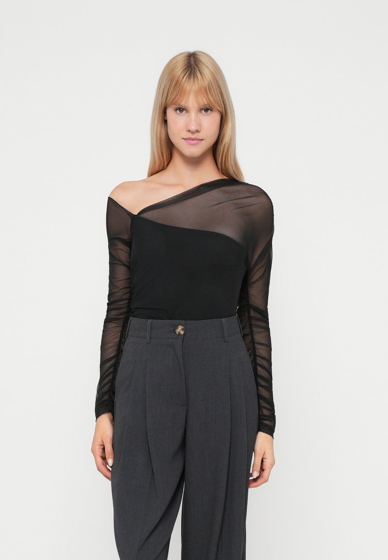 Top lungo a maniche nere in rete con scollatura asimmetrica abbinato a pantaloni grigi a vita alta con pieghe e un bottone decorativo.