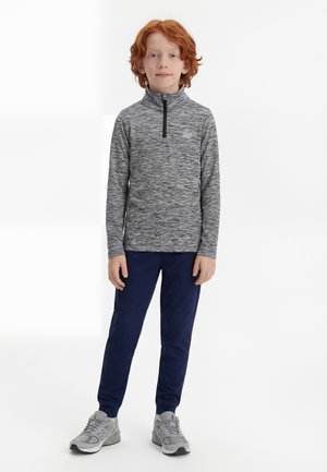 Enfant roux debout portant un pull gris à demi-fermeture éclair, un jogging bleu marine et des baskets grises sur fond blanc.