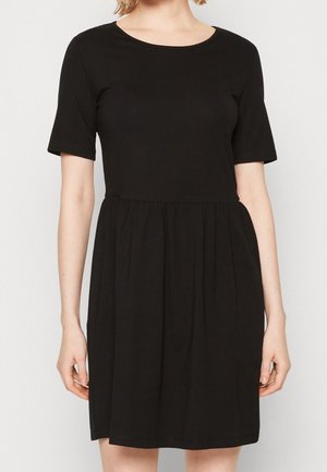 Woman wearing a simple black short-sleeve dress with a gathered waist and knee-length hem against a plain background.
