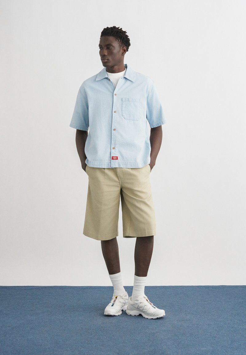 Homme debout les mains dans les poches, portant une chemise à manches courtes bleu clair, un short beige jusqu'aux genoux, des chaussettes blanches et des baskets blanches.