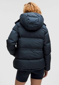 Veste matelassée bleu foncé avec capuche, comportant des sections matelassées et une texture lisse et brillante. Logo au dos ; design ajusté à la taille.