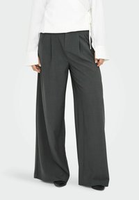 Femme portant un pantalon taille haute, jambe large, gris charbon avec des plis à l'avant, assorti à un haut blanc à manches longues et des chaussures noires.