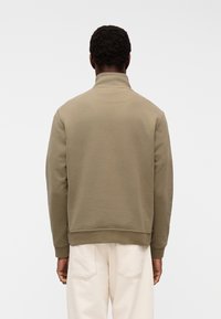 Homme aux cheveux courts et bouclés portant un pull-over uni vert olive à manches longues et un pantalon beige clair, vu de dos.