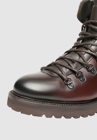 Hnědá kožená turistická bota s lesklým povrchem, kulatou špičkou a robustní gumovou podrážkou. Vybavena pevnými kovovými očními dírkami a hnědými tkaničkami.