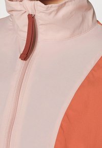 Närbild av en ljusrosa och orange jacka med en framdragkedja och en brun dragkedja på en hög krage.