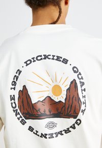 White sweatshirt with a graphic design featuring mountains, a sun, and text “DICKIES QUALITY GARMENTS SINCE 1922.” Bold black and brown colors.
