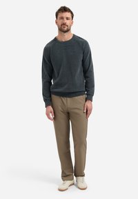 Grijze gebreide trui met een ronde hals, gecombineerd met lichtbruine broek en witte sneakers. Model staat met de handen ontspannen langs de zij.