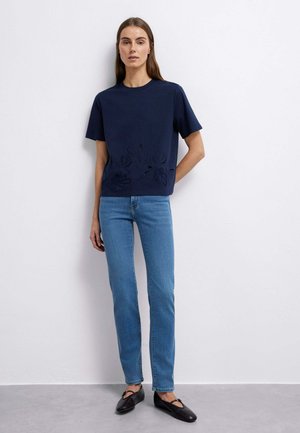 Frau steht vor einer weißen Wand und trägt ein marineblaues, mit Blumen besticktes Kurzarm-Oberteil, hellblaue Jeans und schwarze flache Schuhe.