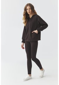 Sudadera con capucha marrón y cierre de cremallera, con puños de canalé, emparejada con leggings a juego y zapatillas blancas. Tejido suave y corte relajado.