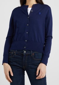 Marinblå cardigan i lättstickad kvalitet med rund halsringning, knappstängning och ribbad fåll. Har en liten broderad logotyp på bröstet.