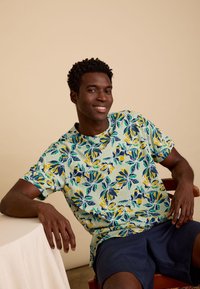 Korte mouwen t-shirt in lichtgroen met een bloemenpatroon featuring blauwe, gele en donkergroene accenten; gecombineerd met marineblauwe shorts.