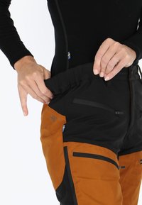 Pantaloni da esterno neri e arancioni con vita elasticizzata, numerose tasche con cerniera e un tessuto resistente e testurizzato.