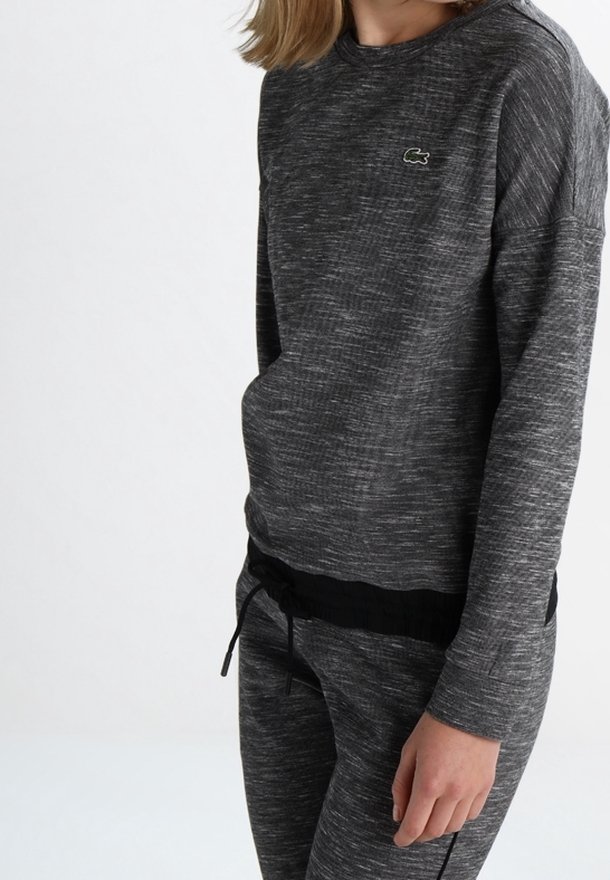 Personne portant un haut gris foncé à manches longues et un pantalon assorti avec un petit logo crocodile sur la poitrine, debout devant un fond blanc uni.