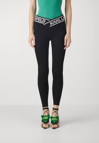 Zwarte leggings met een brede tailleband met daarop "KARL LAGERFELD" in het wit. Gecombineerd met zwarte sandalen met groene fluwelen accenten en enkelbandjes.
