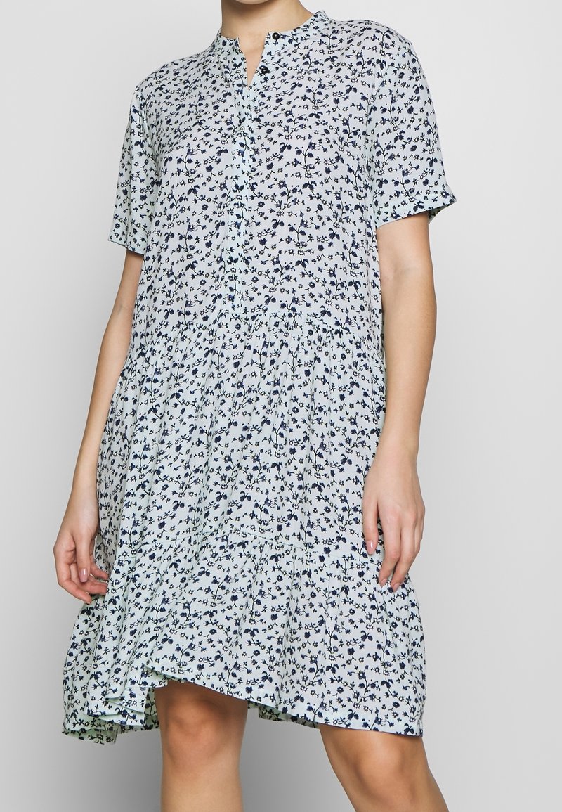 Hellblaues Hemdblusenkleid mit kurzen Ärmeln, versehen mit einem Navy-Blumenmuster. Ausgestellt geschnitten, aus leichtem Stoff gefertigt.
