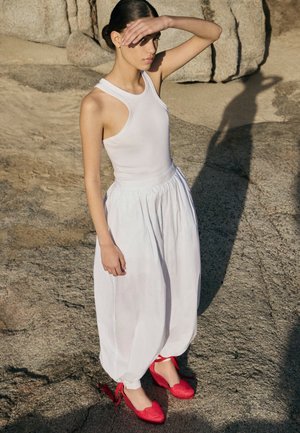 Junge Frau in weißem ärmellosem Top und lockeren weißen Hosen steht auf felsigem Boden, hält die Augen vor der Sonne geschützt und trägt rote Schuhe mit Knöchelriemen.