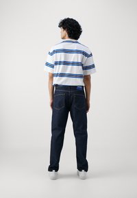Camicetta bianca a maniche corte con righe blu, abbinata a pantaloni jeans blu scuro. I jeans presentano un logo HUGO sulla cintura. Sneakers bianche visibili.