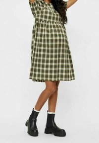 Frau, die ein knielanges grün-weiß kariertes Kleid mit Puffärmeln und schwarze Plattform-Stiefeletten mit weißer Borte trägt.